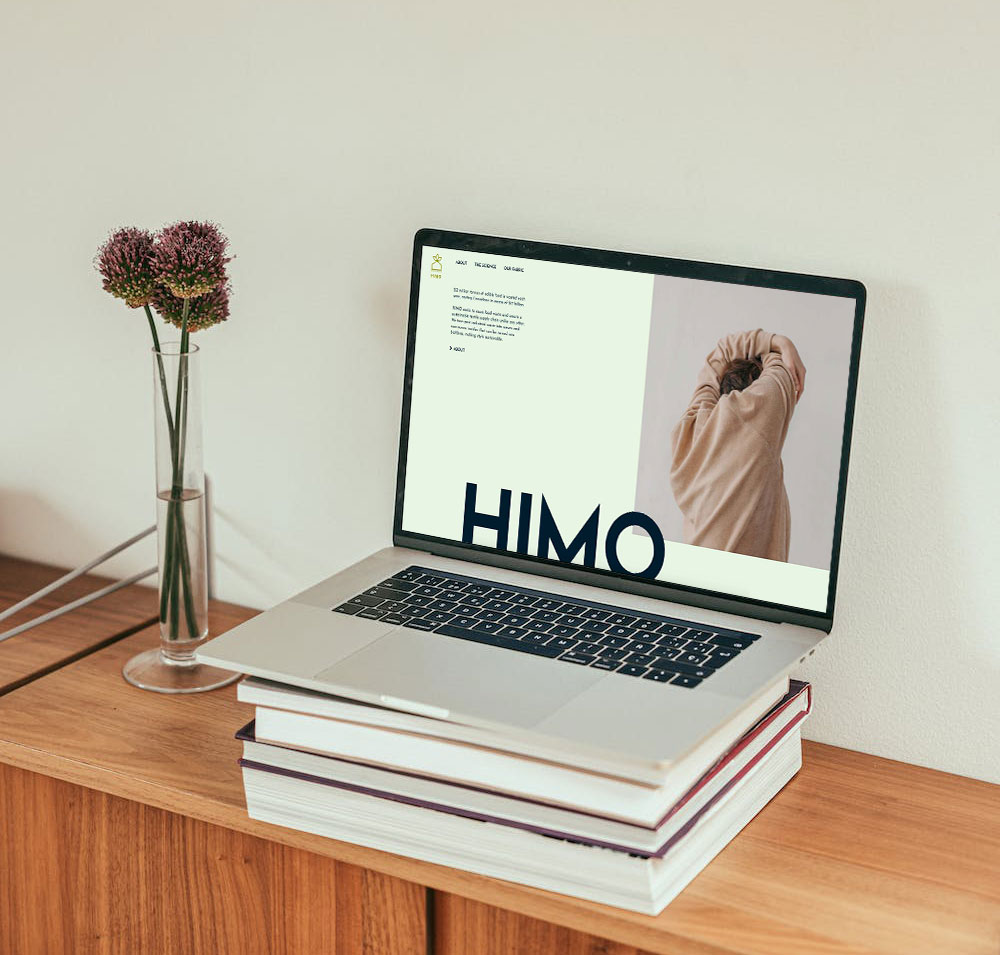 A laptop placed on top of a stack of books showing the home page of HIMO.
